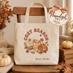 Peut inclure: Un sac fourre-tout en toile couleur crème avec une impression graphique portant le texte COZY SEASON et Your Name. L'illustration comprend un livre ouvert, une tasse de café, des citrouilles, des feuilles d'automne, une bougie et une écharpe.