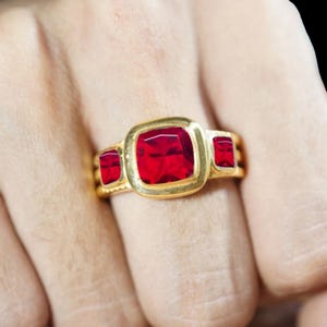 Puede incluir: Un anillo de oro con una gran piedra preciosa roja central de corte cojín, flanqueada por dos piedras rojas más pequeñas de corte rectangular. La banda de metal tiene un acabado liso y pulido con un engaste horizontal para las piedras.