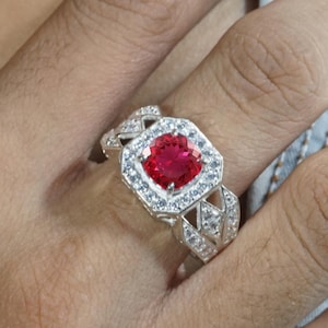 Puede incluir: Un anillo de plata con una piedra central redonda de color rojo vibrante, engastada en un halo rectangular de pequeñas piedras blancas transparentes. La banda presenta un diseño geométrico calado, adornado con numerosas piedras blancas.