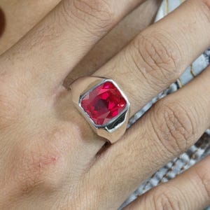 Puede incluir: Un anillo de sello en tono plateado con una gran piedra preciosa roja rectangular facetada en el centro. La banda es lisa y pulida, con una estructura de metal ancha y sólida que se estrecha ligeramente hacia la parte posterior del dedo.