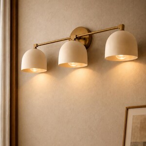 May include: A three-light wall fitting featuring a horizontal brass bar and a circular wall mount. Three matte off-white dome-shaped shades are attached to the bar, each housing a single light bulb that emits a warm, downward-facing glow.