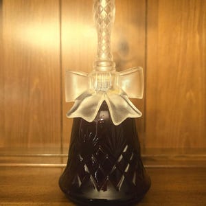May include: A dark purple glass bell featuring a diamond-cut pattern on the body. The handle is clear glass with a textured, cross-hatched design, and a frosted glass bow is positioned at the base of the handle where it meets the bell body.