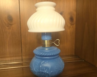 May include: A small oil lamp featuring a blue glass base with embossed floral patterns and a rope-like texture at the neck. The lamp includes a gold-colored metal burner with a heart-shaped adjustment knob and a white, ribbed glass shade.