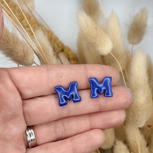 May include: A pair of stud earrings shaped like the letter M, featuring a glossy, deep blue finish with a slightly textured, rounded surface. The letter M is rendered in a bold, block-style font with distinct serifs, creating a uniform appearance.