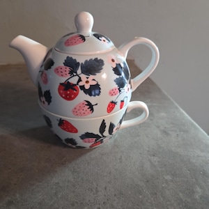 May include: A ceramic tea-for-one set featuring a light blue teapot stacked on a matching teacup. The set is decorated with a repeating pattern of red and pink strawberries, dark green leaves, and small white flowers across the entire surface.