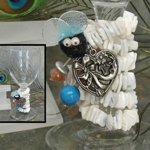 May include: A glass wine charm with a white heart charm, a blue and silver butterfly charm, and white beads. The charm is attached to a glass wine glass.