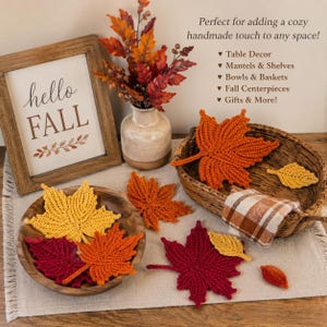 May include: A collection of crocheted maple leaves in yellow, orange, and deep red. The leaves are arranged in a wooden bowl and a woven basket alongside a framed sign that reads hello FALL, and a vase filled with artificial autumn foliage.