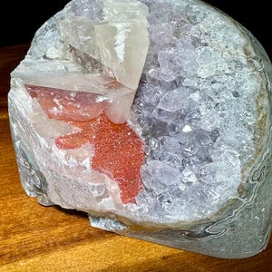May include: A geode specimen featuring a cluster of translucent, pale purple amethyst crystals on one side. The other side displays large, clear, angular calcite crystals layered over a vibrant, textured patch of reddish-orange hematite inclusions.