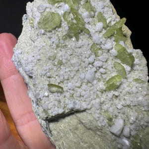 May include: A mineral specimen featuring a light green, textured rock matrix covered in clusters of small, translucent white crystals and elongated, pale olive-green crystalline formations scattered across the surface.