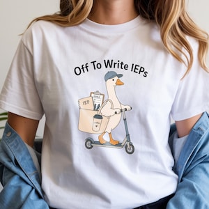 May include: A white t-shirt featuring a graphic of a goose wearing a blue cap and riding a scooter. The goose carries a beige tote bag containing a notebook labeled IEP, a clipboard labeled GOALS, and a coffee cup. The text reads Off To Write IEPs.