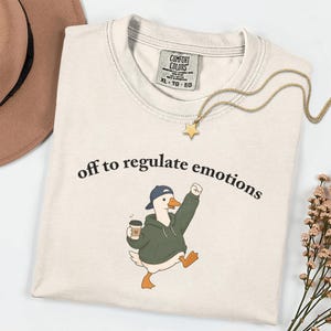 Puede incluir: Una camiseta de cuello redondo color beige con el gráfico de un ganso blanco que lleva una sudadera con capucha verde oscuro y una gorra azul, sosteniendo un café. El texto "off to regulate emotions" está impreso en negro sobre el ganso. Etiqueta Comfort Colors.