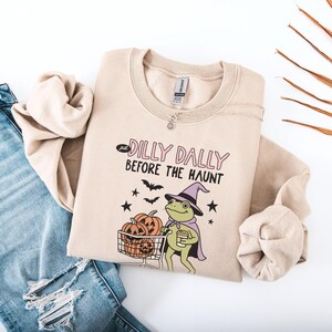 Puede incluir: Una sudadera beige de cuello redondo con un gráfico de una rana verde con sombrero y capa de bruja morada, empujando un carrito de la compra lleno de calabazas naranjas. El texto dice "just DILLY DALLY BEFORE THE HAUNT" con murciélagos y estrellas.