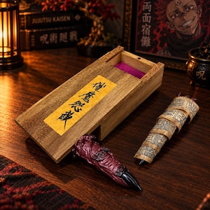 May include: A wooden box with a sliding lid featuring a yellow label with black Japanese text, containing a purple velvet lining. Beside the box lies a textured, dark red finger-like object with a black tip and a tan scroll with black patterns.