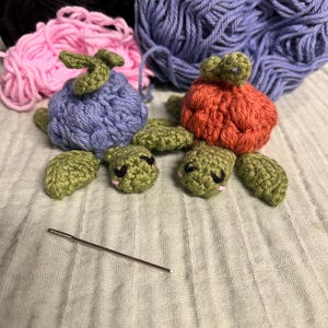 Può includere: Due piccole tartarughe all'uncinetto con teste e pinne verde oliva. Una tartaruga ha un guscio viola testurizzato, l'altra un guscio arancione. Entrambe presentano occhi neri ricamati e piccole guance rosa. Un ago da cucito è posizionato nelle vicinanze.
