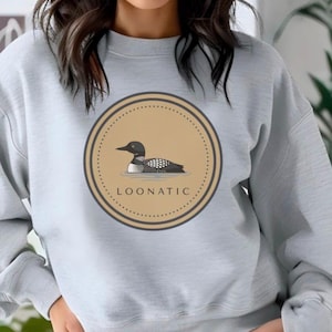 May include: A light gray crewneck sweatshirt featuring a circular graphic on the chest. The graphic contains a tan background with a black and white loon illustration and the text LOONATIC printed in black capital letters inside the border.