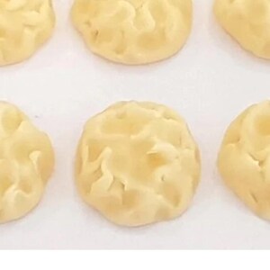 Peut inclure: Six biscuits ronds, de couleur jaune pâle, disposés en deux rangées de trois. Chaque biscuit présente une surface texturée, bosselée et irrégulière avec de petites indentations, offrant un aspect uniforme sur toutes les pièces.
