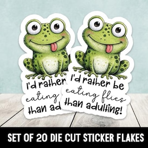 May include: Two die-cut stickers featuring a cartoon frog with big eyes and a pink tongue sticking out. The frog is green with dark green spots. Text on the stickers reads "I'd rather be eating flies than adulting!"