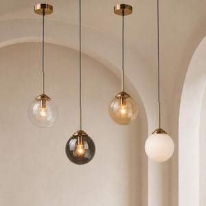 May include: Four pendant lights hanging from the ceiling, each featuring a gold metal fixture and a spherical shade. The shades include one clear glass, one dark grey glass, one amber glass, and one opaque white glass, all suspended by black cords.