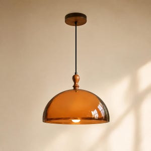 Peut inclure: Une suspension lumineuse dotée d'un grand abat-jour en verre ambré en forme de dôme. Le luminaire comprend un embout en bois brun foncé au sommet, un cordon noir fin et une rosace de plafond circulaire brun foncé. Une ampoule est visible à l'intérieur.