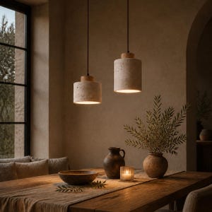 Op de afbeelding: Twee cilindrische hanglampen met getextureerde beige stenen kappen en houten bovenkanten, opgehangen aan zwarte snoeren. De lampen hangen boven een houten tafel met een donkere keramische kan, een glazen kaarshouder en een ronde keramische vaas.