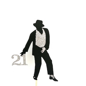 May include: Black silhouette of a person wearing a hat and a suit, holding a silver glitter number 21. The figure is on a wooden stick.