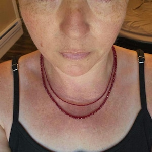 May include: A double-strand necklace featuring small, faceted, deep red gemstone beads. The necklace rests against the neck, with the two strands creating a layered effect. The beads are uniform in size and color, reflecting light across their surfaces.