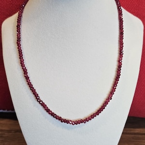 May include: A single strand necklace composed of small, round, faceted deep red garnet beads. The beads are uniform in size and strung together to form a continuous circular chain that rests evenly around the neck of the display form.