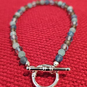 May include: A beaded bracelet featuring a mix of faceted and smooth round beads in shades of blue, green, and brown. The bracelet is secured with a polished silver-toned toggle clasp consisting of a circular ring and a horizontal bar component.