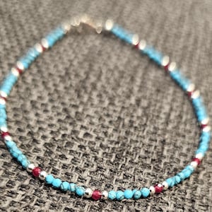 May include: A delicate beaded bracelet featuring small, faceted turquoise-colored stones interspersed with polished silver-toned beads and vibrant, deep red accent beads arranged in a repeating pattern along the entire length of the circular strand.