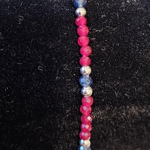 May include: A beaded bracelet featuring a repeating pattern of faceted hot pink beads, smooth metallic silver spheres, and faceted dark blue beads. The small, round beads are strung together in a linear arrangement, creating a colorful accessory.