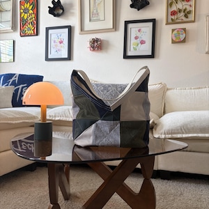 May include: A patchwork tote bag constructed from various fabric panels in shades of olive green, navy blue, and white with pinstriped patterns. The bag features a wide, integrated shoulder strap and a structured, multi-paneled design with visible stitching.