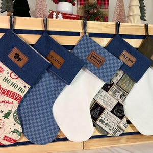 May include: Five Christmas stockings hanging in a row. The first features a cream fabric with red and green holiday text and Santa graphics. The second is blue denim with a checkered pattern. The third is white with a blue checkered cuff. The fourth has a plaid pattern. The fifth is solid white with an olive green cuff. Each stocking has a brown leather name patch.