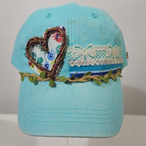 May include: A light blue baseball cap featuring a heart-shaped twig frame over a floral fabric patch, a horizontal strip of white lace trim, and a decorative twine vine with small green leaves wrapped around the base of the crown.