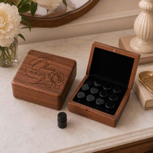 Puede incluir: Una caja de almacenamiento de madera oscura con tapa abatible grabada con el nombre Olivia, la fecha 18.06.2024 y un dibujo floral. La caja abierta contiene un inserto de terciopelo negro con doce pequeños frascos cilíndricos negros.