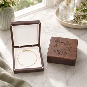 May include: A dark wood jewellery box with a white interior lining containing a thin gold chain bracelet. The lid features an engraved hummingbird, a flower, the name GRACE, and the date 12.20.2026. The box is square with metal hinges.