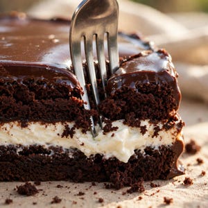 Puede incluir: Una porción de pastel de chocolate con una capa gruesa de relleno de crema blanca entre dos capas de bizcocho de chocolate oscuro, cubierto con un glaseado de ganache de chocolate suave. Un tenedor de metal plateado está insertado.