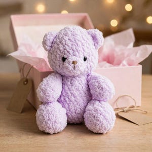 Puede incluir: Un oso de peluche hecho de hilo suave de color morado claro con una apariencia texturizada y difusa. El oso presenta pequeños ojos negros y una nariz marrón cosida. Está colocado frente a una caja de regalo rosa claro abierta.