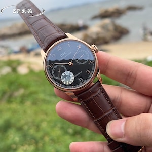 May include: An IWC Schaffhausen automatic wristwatch with a round rose gold case, a black dial featuring Arabic numerals, a date window at the six o'clock position, two sub-dials, and a dark brown alligator-textured leather strap with a buckle.