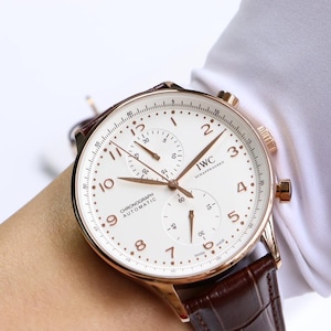 May include: An IWC Schaffhausen chronograph automatic watch with a rose gold-tone case, a white dial featuring Arabic numerals, two sub-dials, and thin hands. The timepiece is attached to a dark brown leather strap with a textured crocodile pattern.