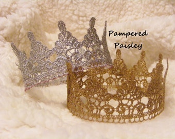 May include: Two decorative lace crowns, one silver and one gold, sit on a soft, white, textured surface. The gold crown is in the foreground, with the silver crown behind it. The text "Pampered Paisley" is visible.