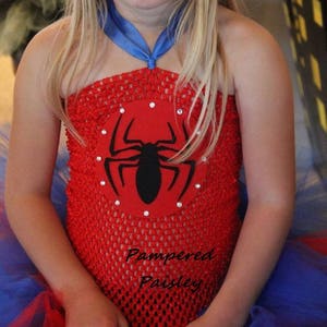 May include: A red crochet tube top featuring a circular red felt patch with a black spider graphic and small clear rhinestones. The top includes a blue ribbon halter strap and is layered over a blue and red tulle skirt. Text reads Pampered Paisley.