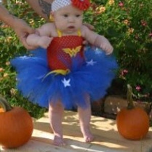 May include: A baby outfit featuring a red bodice with a gold Wonder Woman logo, a blue tulle tutu skirt adorned with white stars, and a white headband with a large red flower. The ensemble is designed for an infant and includes a matching top.