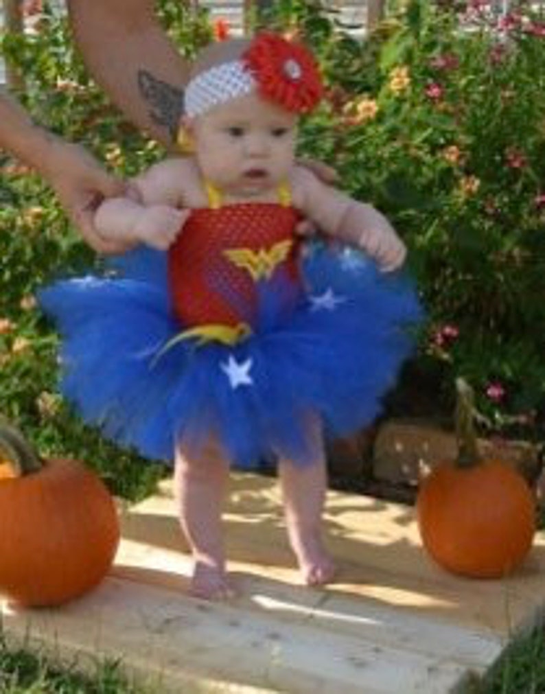 May include: A baby outfit featuring a red bodice with a gold Wonder Woman logo, a blue tulle tutu skirt adorned with white stars, and a white headband with a large red flower. The ensemble is designed for an infant and includes a matching top.