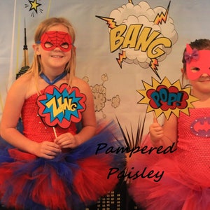 May include: Two superhero costumes featuring red and pink tube tops with matching tulle skirts. One costume includes a red spider-web mask and a ZING sign, while the other has a pink bat-style mask and a POP sign. The text Pampered Poisley is visible.