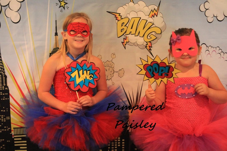May include: Two superhero costumes featuring red and pink tube tops with matching tulle skirts. One costume includes a red spider-web mask and a ZING sign, while the other has a pink bat-style mask and a POP sign. The text Pampered Poisley is visible.