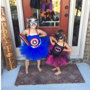 May include: Two children's costumes: one blue Captain America dress with a star emblem, matching mask, and blue tulle skirt; the other a black Batman dress with a pink bat logo, matching mask, and a layered black and pink tulle skirt.