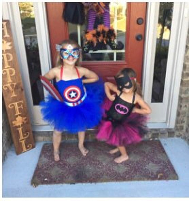 May include: Two children's costumes: one blue Captain America dress with a star emblem, matching mask, and blue tulle skirt; the other a black Batman dress with a pink bat logo, matching mask, and a layered black and pink tulle skirt.