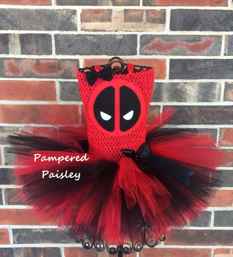 K&ouml;nnte beinhalten: Ein rotes H&auml;kelkleid mit einem schwarzen Deadpool-Masken-Emblem auf der Brust, einer kleinen schwarzen Schleife am Ausschnitt und einem mehrlagigen T&uuml;llrock in abwechselnd roten und schwarzen Farben. Der Text Pampered Paisley ist links zu sehen.