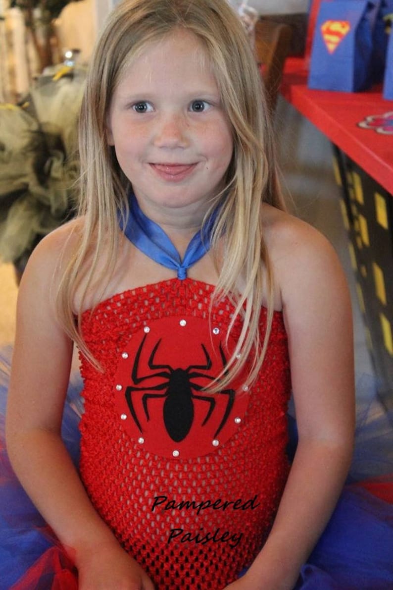 May include: A red crochet tube top featuring a circular red felt patch with a black spider graphic and small clear rhinestones. The top includes a blue ribbon halter strap and is layered over a blue and red tulle skirt. Text reads Pampered Paisley.