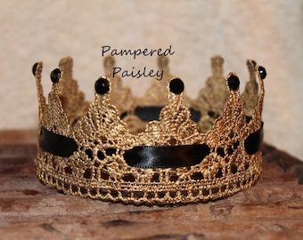 May include: A gold crocheted crown featuring a black ribbon woven through the base and small black beads at each point. The text Pampered Paisley is visible above the crown. The crown has a detailed lace-like texture and a circular shape.